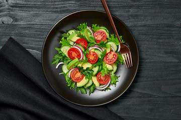 Fresh salad with cherry tomatoes, avocado,cucumeber and lettuce on dish. Healthy vegetarian meal concept. Tasty mixed leaves, tomatoes and mix vegetable salad on black wood, top view.