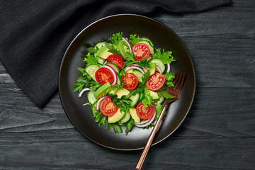 Fresh salad with cherry tomatoes, avocado,cucumeber and lettuce on dish. Healthy vegetarian meal concept. Tasty mixed leaves, tomatoes and mix vegetable salad on black wood, top view.