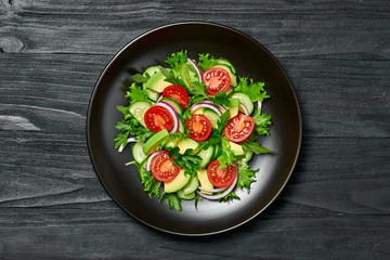Fresh salad with cherry tomatoes, avocado,cucumeber and lettuce on dish. Healthy vegetarian meal concept. Tasty mixed leaves, tomatoes and mix vegetable salad on black wood, top view.