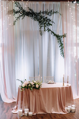 Wedding presidium in restaurant, free space. Wedding banquet table for newlyweds with flowers, greenery, pink cloth and bulbs. Lush floral arrangement on wedding table. Luxury wedding decorations