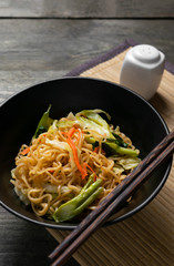 Fried instant noodle with carrot and kale in black bowl on wooden table.