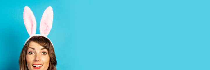 Young woman with rabbit ears looks out over a blue background. Easter concept, surprise, hide and seek. Banner