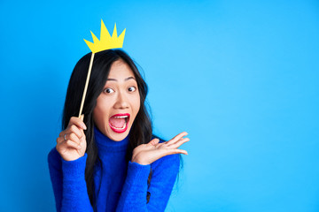 Emotional suprised woman with paper crown on stick on blue background