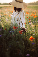 Stylish girl in hat walking in wildflowers in sunset light in summer meadow. Young woman in linen...