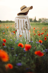 Young woman in rustic linen dress walking among poppy and cornflowers in summer meadow countryside...