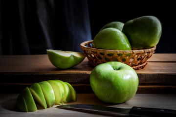 Still life of green apples lying in a basket and on boards, on a black background with light illumination, shallow depth of field. Conception, healthy food.