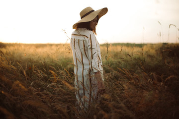 Woman in rustic dress and hat walking in wildflowers and herbs in sunset golden light in summer meadow. Atmospheric authentic moment. Stylish girl enjoying evening in countryside.