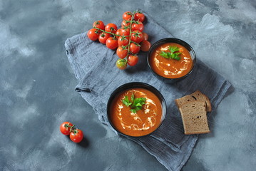 Hot winter soup. Red hot tomato soup with garlic, sweet paprika, parsley, served with cream and sourdough bread in two black ceramic bowls on a gray background