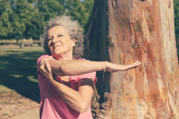 Happy old lady warming up for morning exercise in park. Senior grey haired woman in casual sitting on bench and stretching hand. Age and fitness concept