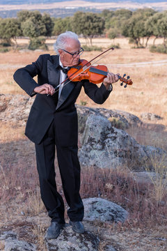 Elderly man, dressed in tuxedo, plays the violin in a natural environment