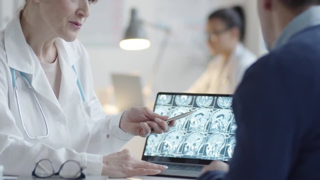 Tilt up shot of middle aged female doctor zooming x-ray image of brain on laptop screen and explaining diagnosis to male patient during consultation in medical office
