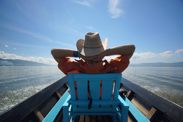 Young man in hat sitting on chair in boat with hands behind head and enjoying life, blue sky and...