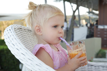 Happy toddler girl sitting at the restaurant table and drinking fresh-squeezed orange juice	