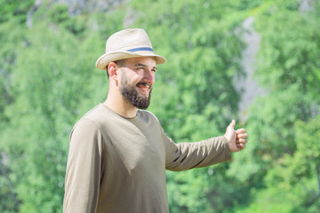 Bearded man in a baseball cap, in a T-shirt and jeans on a background of nature, smiles and votes. hitchhiking travel concept