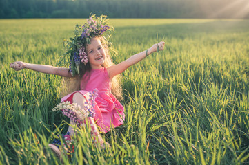 Naklejka na ściany i meble Beautiful little girl in a pink dress and boots with a wreath of flowers on her head sits on the green grass