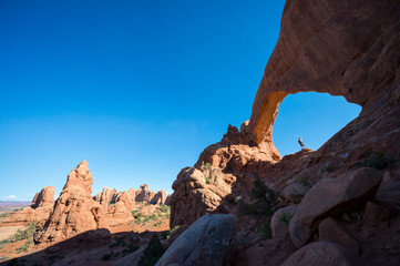 Distant view of hiker standing under a dramatic natural rock arch in red rock desert landscape