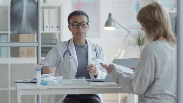 Mixed raced female physician in lab coat and glasses speaking with woman and giving medical service agreement to her for signing while having consultation in clinic