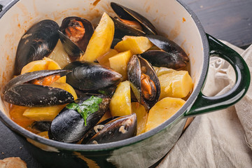 Close-up mussels and fried potatoes in sweet and sour sauce in a saucepan and chabatta on a dark wooden table. Mediterranean traditional cuisine.