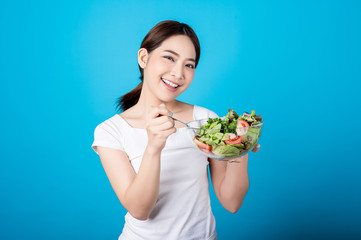 Beautiful young asian eating salad with big smile happy beaming face in seamless blue isolated background. Diet healthy concept. Her face and skin are healthy, fresh, bright and youthful.