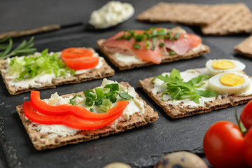 Crispbread with tasty cream cheese, salmon, eggs and vegetables on dark background, closeup