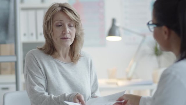 Tilt down shot of cheerful middle aged blonde woman shaking hands with female doctor at desk, giving her medical history and discussing healthcare problems in clinic