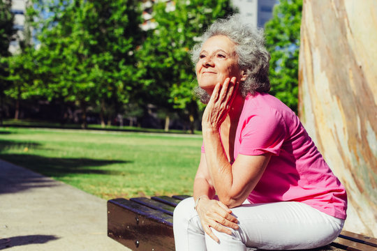 Joyful old lady waiting someone in city park. Senior grey haired woman in casual sitting on bench outdoors, leaning chin on hand and looking into distance. City park concept