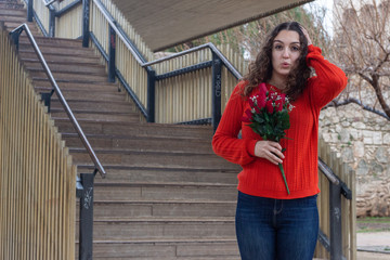 Portrait of attractive caucasian young woman with roses being surprised, opening her eyes and mouth with shock, touching her head with hands, in the park, orange sweater and jeans, long curly hair. Pl