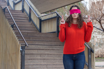 Portrait of attractive caucasian young woman with eye tape keeping finger crossed, having calm expression, in the park, orange sweater and jeans, long curly hair. Place for your text in copy space.