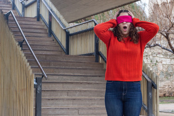 Portrait of attractive caucasian young woman with eye tape being surprised, opening her eyes and mouth with shock, touching her head with hands, in the park, orange sweater and jeans, long curly hair.