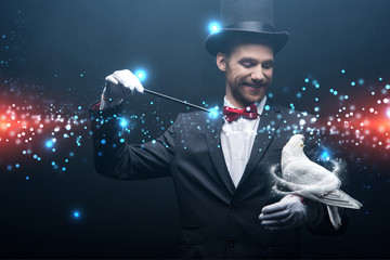 happy magician in hat making abracadabra with dove and wand in dark room with smoke and glowing...