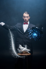 shocked magician showing trick with dove, wand and hat in dark room with smoke and glowing...