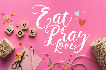 Obraz na płótnie Canvas top view of valentines decoration, scissors, gift box, twine and love lettering on wooden cubes on pink background with eat, pray, love lettering