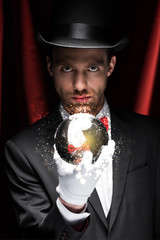 professional magician holding magic ball with glowing illustration in circus with red curtains