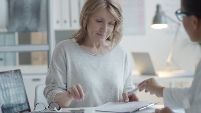 Middle aged blonde woman taking clipboard from female doctor, putting on glasses and signing medical service agreement during healthcare consultation