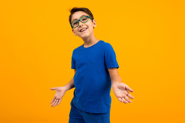 handsome teenager boy in a blue t-shirt with glasses spreads his hands on a yellow background with...