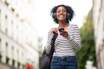 smiling african american woman walking with bag and mobile phone listening to music with headphones in city