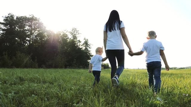 happy family teamwork mother, little brother and sister walk in the park nature holding hands slow motion video concept. mom, kids boy and girl daughter and son hold hands go on green grass sun