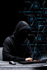hacker in mask sitting near computer monitor and typing on computer keyboard near padlocks on black