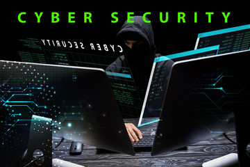 hacker in hood sitting near computer monitors near cyber security lettering on black