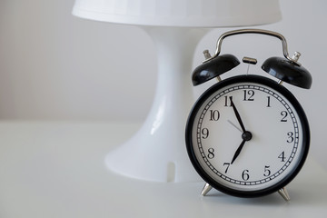A black alarm clock stands on a white nightstand against a white lamp and white wall. Early rise....