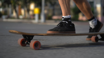 The legs of a light-skinned man on a longboard. Men's feet in dark shoes move along a paved city...