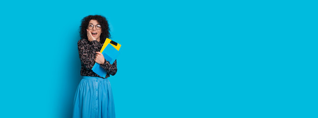 Cheerful caucasian student with curly hair surprised by something while posing on a blue wall with free space