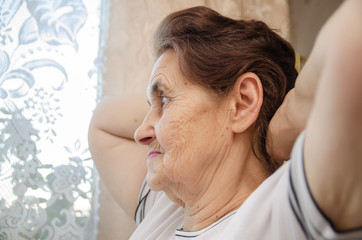 elderly dark haired woman with her hands behind her head looks out the window