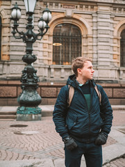 Fototapeta na wymiar Portrait of a young blond man in a jacket with a backpack, stands against the background of a beautiful old building with columns, a lantern in St. Petersburg