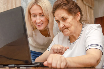 Smiling young blonde girl teaches an elderly dark-haired woman, grandmother how to work on a laptop at home