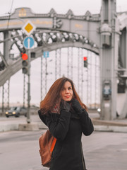 Obraz premium Portrait of a beautiful brunette girl with long hair in a coat and with a bright scarf, stands against the background of the river with a bridge, on the bridge busy traffic