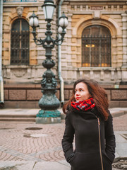 Portrait of a beautiful brunette girl with long hair in a coat and a bright scarf, stands against the background of a beautiful old building with columns, a lantern in St. Petersburg