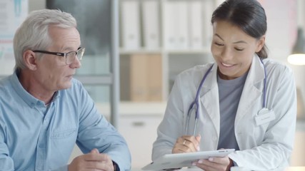 Tracking shot of young mixed raced female doctor showing medical test results on digital tablet and talking with senior caucasian man during consultation - Powered by Adobe