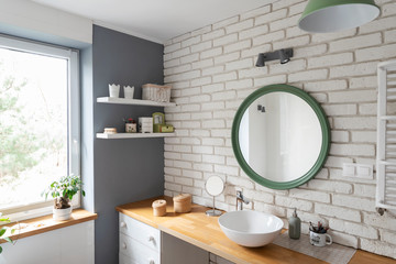 interior of modern bathroom with white brick wall, window and round mirror. Wooden furniture and counter in design indoor.
