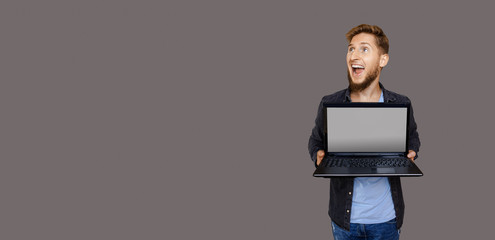Ginger caucasian man with beard is looking up to gray blank space while holding a laptop and...